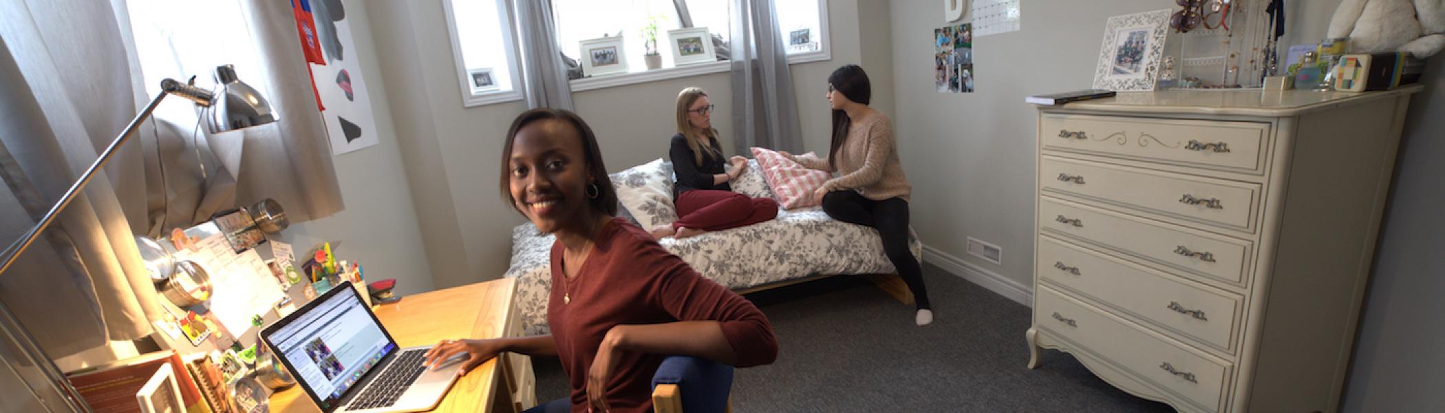 A girl sitting at her desk in her dorm room smiling with two girls sitting on her bed