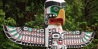 The opv of an indigenous totem pole outside in the summertime set against a wall of evergreen trees