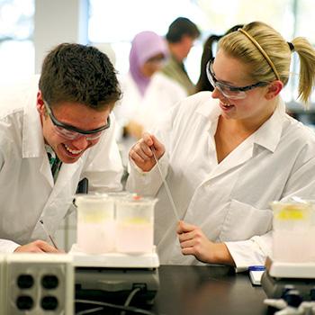2 Students standing side by side in a chemistry lab in their lab coats with windows in the background