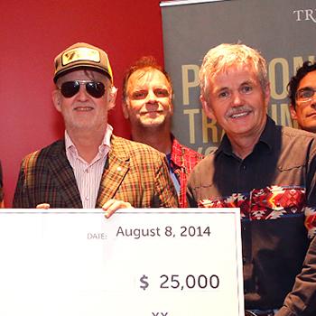 Dr. Leo Groarke, Julie Davis and Lee Hays posing with a large white check for $25,000 with the band Blue Rodeo in front of a red wall